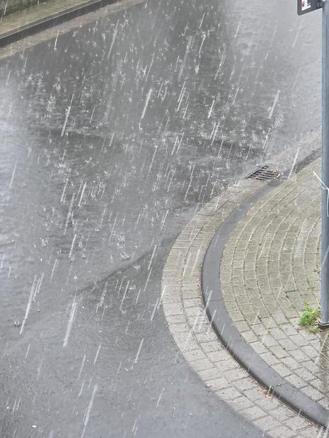 Foto zeigt große Regentropfen, die auf eine Straße und einen Gehweg fallen. (Quelle DWD - Deutscher Wetterdienst)